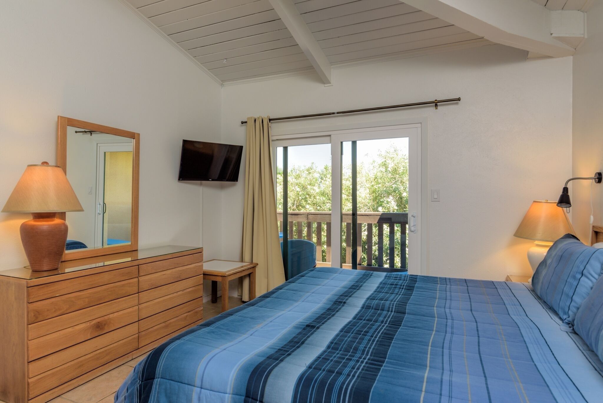 A bedroom with a bed, a dresser, a television, and a balcony with a view of trees.
