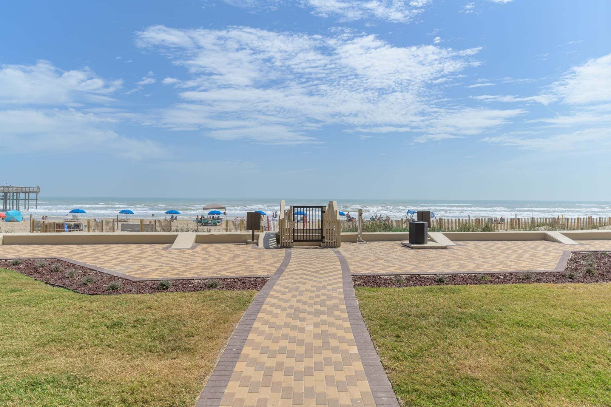 Un paseo marítimo frente a la playa que conduce a una entrada con portón, sombrillas y una terraza de madera.