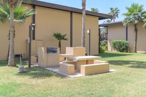 Área de varanda externa com mesa e bancos de concreto, cercada por palmeiras e um edifício com fachada bege.