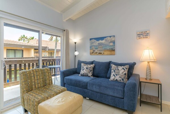 Uma sala de estar com um sofá azul, uma poltrona estampada e uma mesinha com abajur. Há um quadro com uma cena de praia na parede e um letreiro escrito 'I AM AN AQUAHOLIC LET'S GET SHIP FACED!'