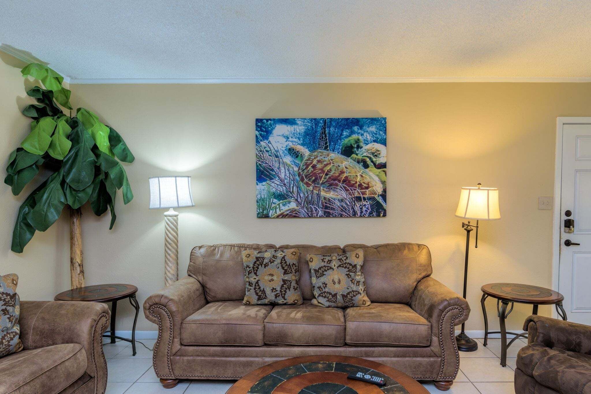 Ein Wohnzimmer mit braunem Sofa, zwei Beistelltischen, einem Couchtisch, einer großen Grünpflanze und einem Bild an der Wand.