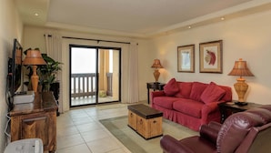 Un salón con un sofá rojo, una mesa de centro de madera y un televisor sobre un mueble de madera. Hay una puerta corrediza de vidrio que da a un balcón.