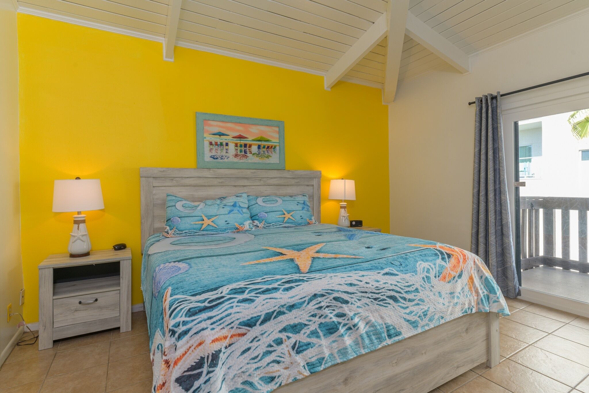 Un dormitorio con una pared de color amarillo brillante, una cama con colcha de temática de estrella de mar, dos lámparas de noche y un cuadro sobre la cama.