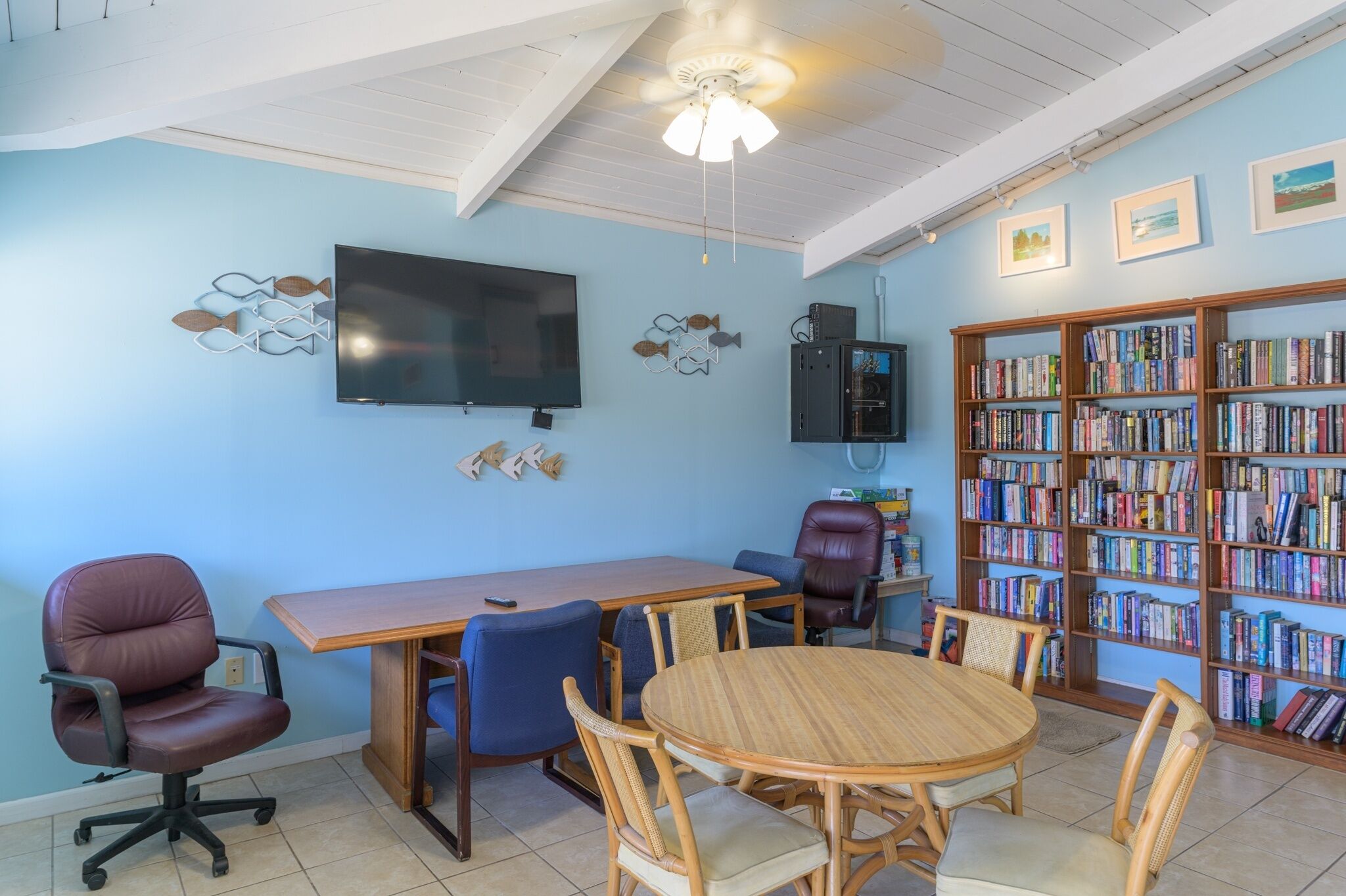 A room with a wooden table and chairs, a television, a bookshelf, and a ceiling fan.