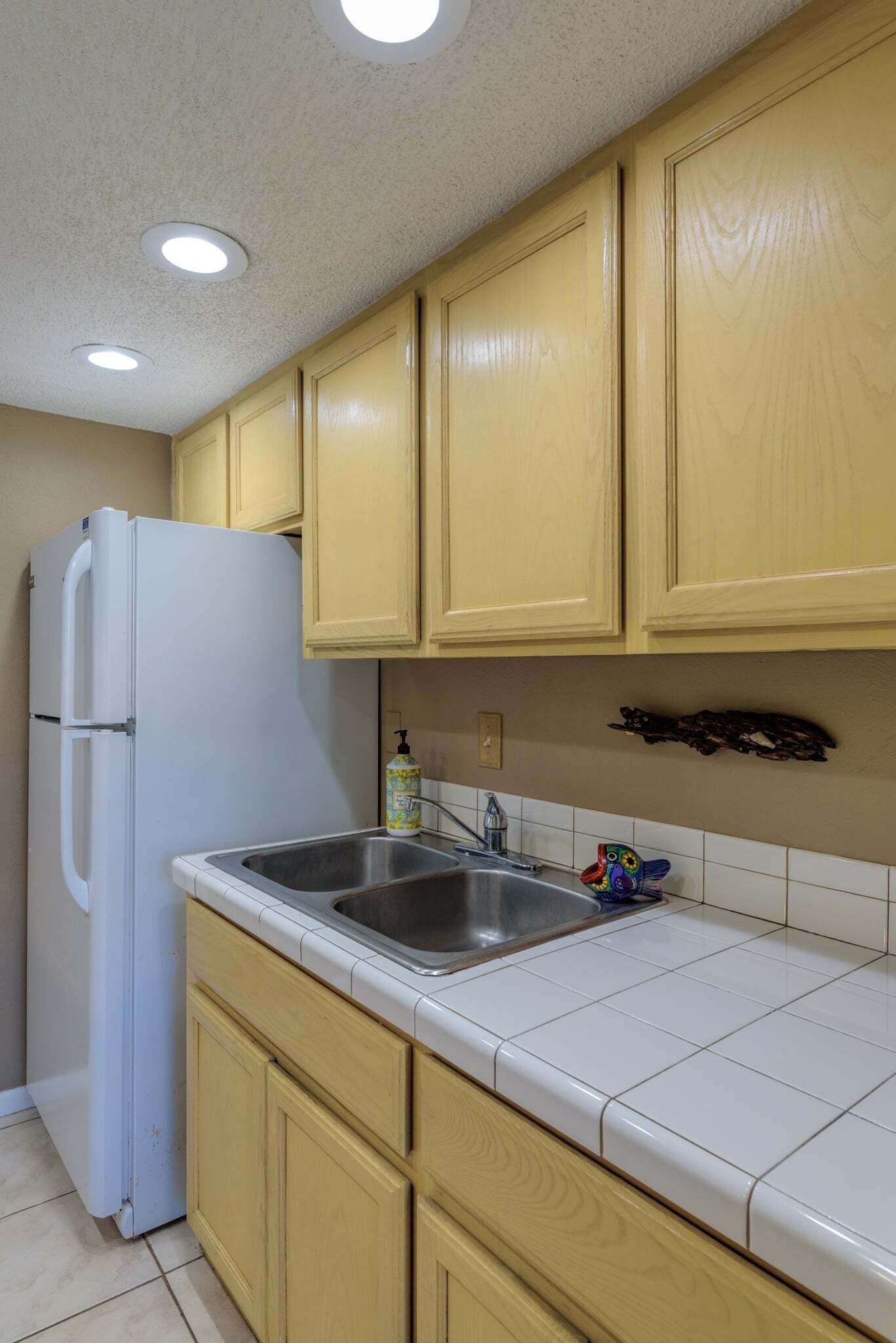 A kitchen with wooden cabinets, a white refrigerator, a stainless steel sink, and a tiled backsplash.