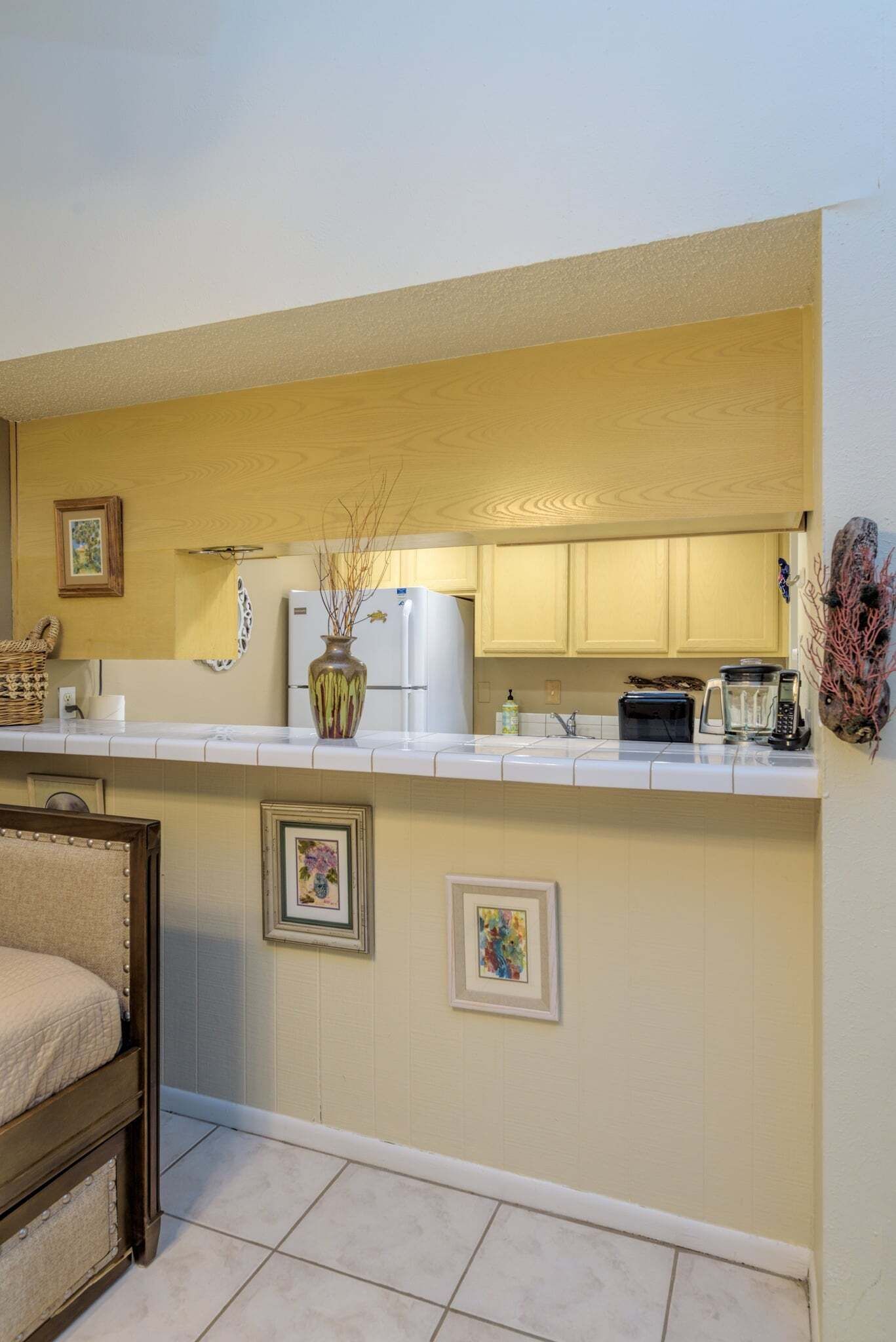 A compact kitchen with white cabinets, a refrigerator, a coffee maker, and a toaster. There are framed pictures on the wall and a small table with a chair.