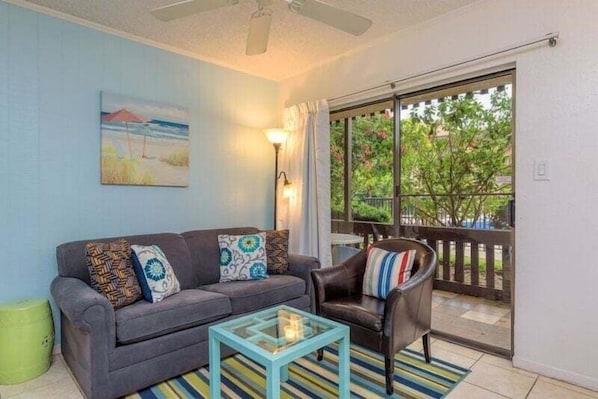A living room with a sofa, a coffee table, a lamp, and a view of the outdoors through a sliding door. - Tiki 111 - Ocean View Condo In South Padre Island 1 Bedroom Condo (South Padre Island)