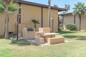 An outdoor patio area with a concrete table and benches, surrounded by palm trees and a building with a beige exterior. - Tiki 111 - Ocean View Condo In South Padre Island 1 Bedroom Condo (South Padre Island)