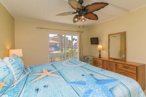 A bedroom with a bed, a ceiling fan, a dresser, a mirror, and a view of a building outside. - Tiki 158 - Standard 1BR Beach Area Condo (South Padre Island)