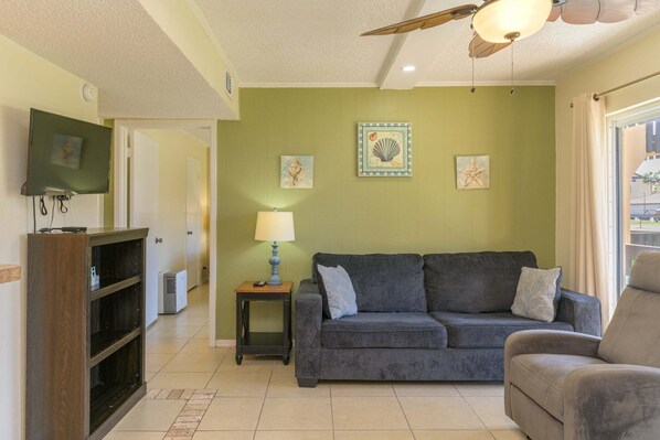 A living room with a green wall, a grey sofa, a wooden cabinet, and a ceiling fan. - Tiki 158 - Standard 1BR Beach Area Condo (South Padre Island)