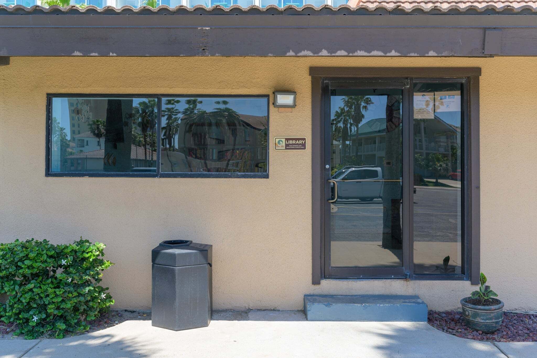 A building with a beige exterior, a glass door entrance, a trash bin, and a small potted plant.