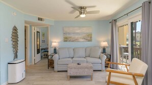 Ein Wohnzimmer mit grauem Sofa, einem Holzstuhl, einem Deckenventilator und einem Strandbild.