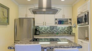 Una cocina con armarios blancos, un refrigerador de acero inoxidable, un microondas, una isla de vidrio con dos sillas y un salpicadero de azulejos.