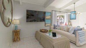 A living room with a beige sofa, a coffee table with a plant, a large mirror, and a painting of a beach scene. - Tiki 325 - Family Friendly Oceanview Condo Sleep 6 1 Bedroom Condo (South Padre Island)