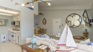 A modern kitchen with white appliances, a dining area with a wooden table and chairs, and a living room with a sailboat model. - Tiki 325 - Family Friendly Oceanview Condo Sleep 6 1 Bedroom Condo (South Padre Island)