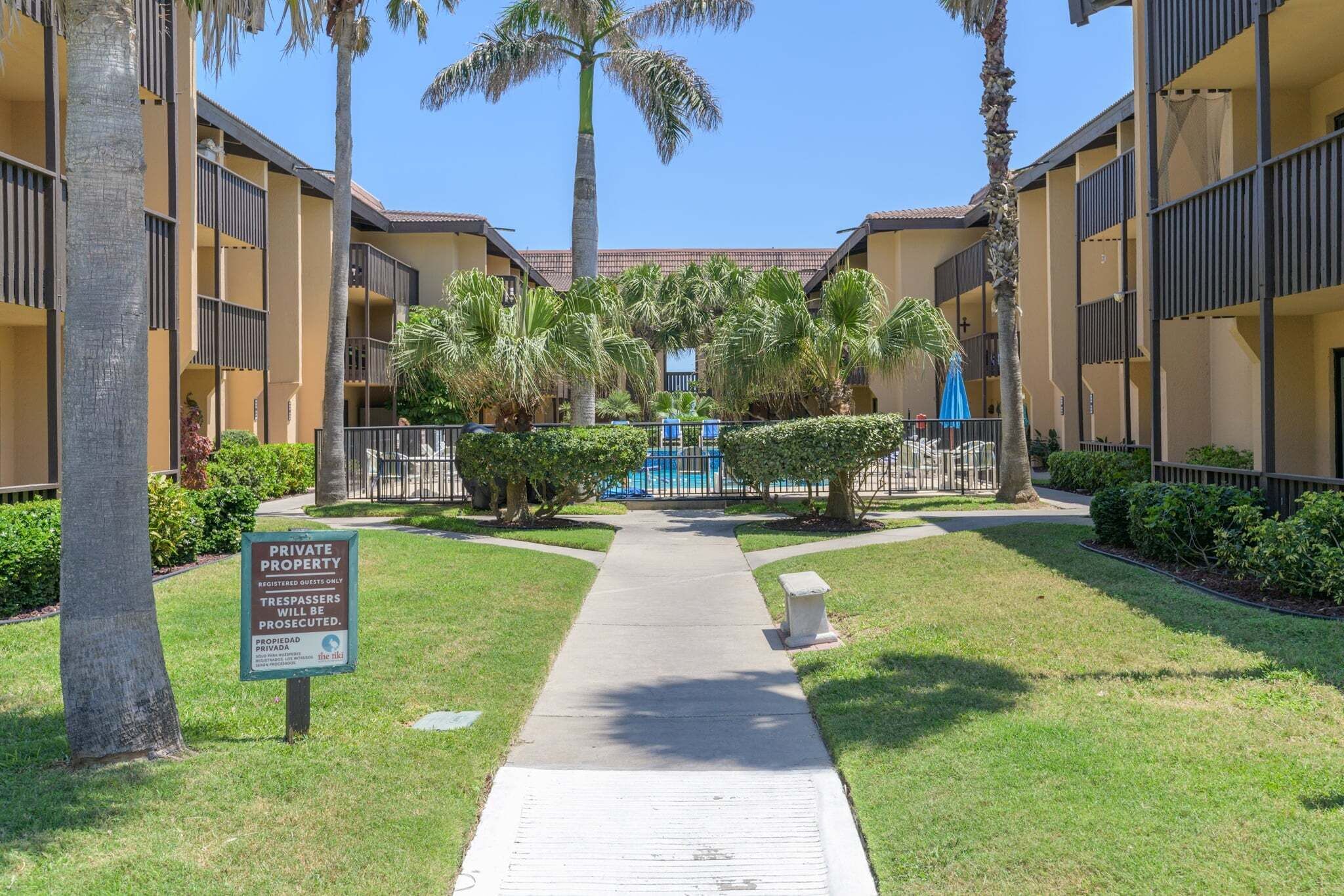A private property with a pathway leading to a building with balconies, surrounded by palm trees and greenery.