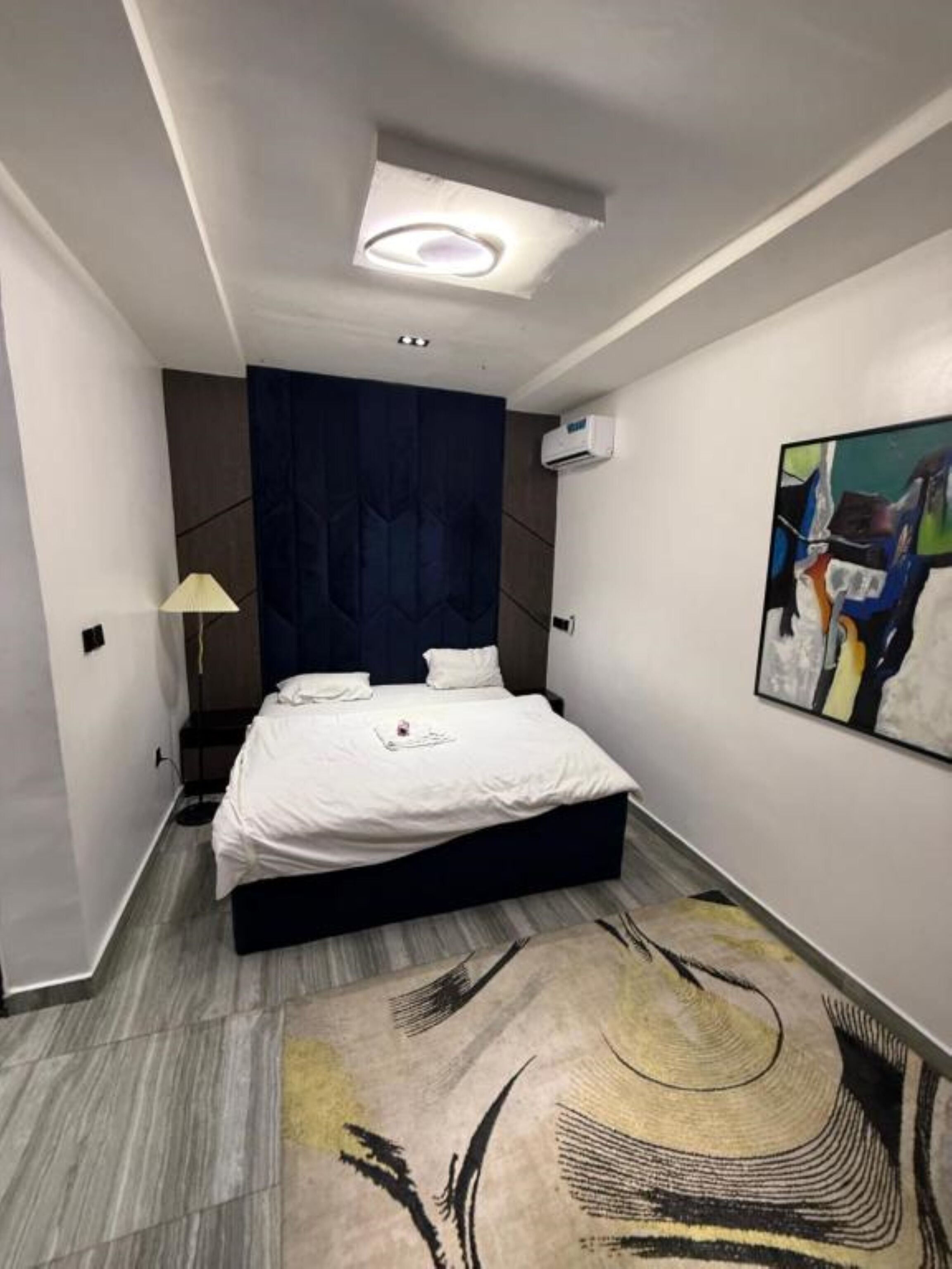 Een moderne slaapkamer met een bed, een hoofdbord, een vloerlamp, een schilderij en een plafondlamp.