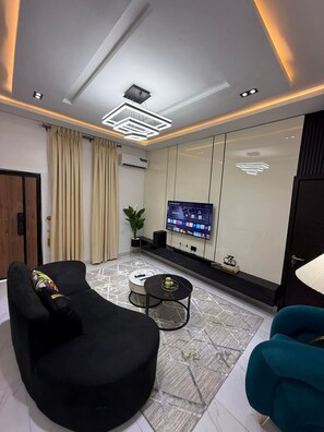 A modern living room with a black sofa, a teal armchair, a coffee table, and a flat-screen TV mounted on the wall. - Muzah Homes - Port Harcourt (Port Harcourt)