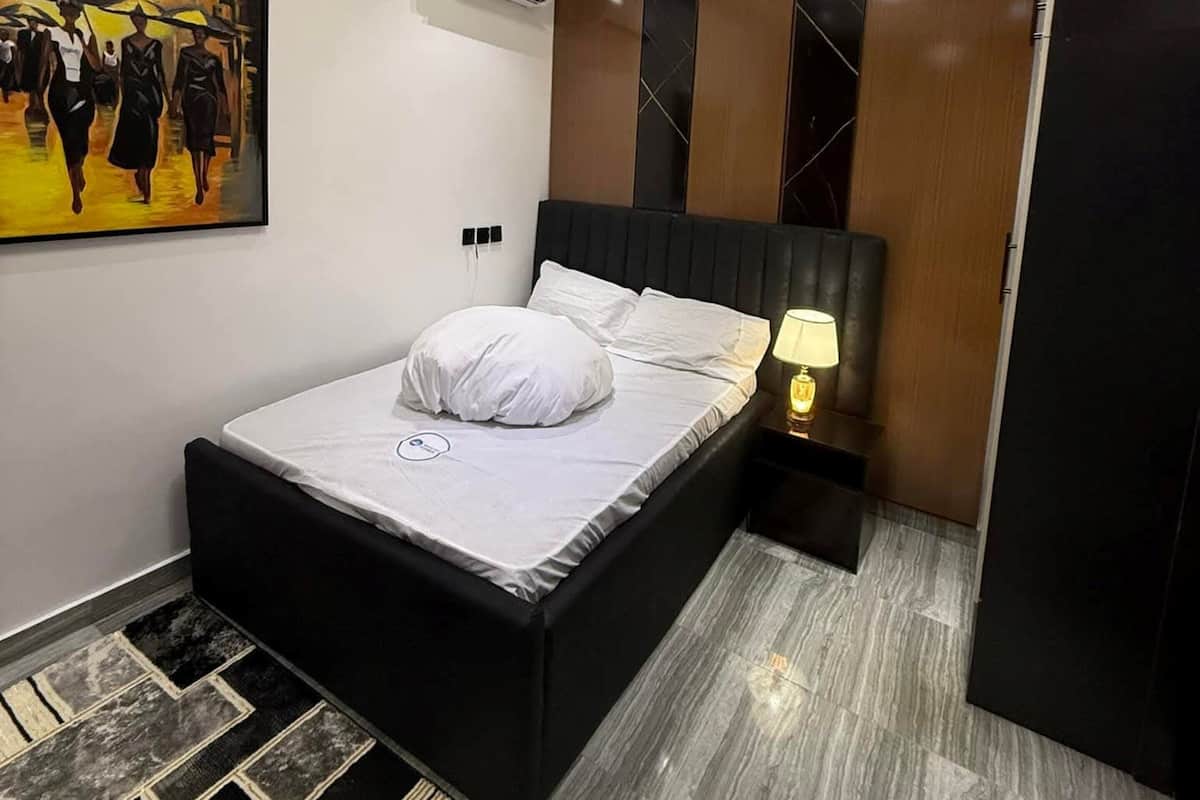 Un dormitorio moderno con una cama, una mesita de noche, una lámpara, un cuadro y un aire acondicionado.