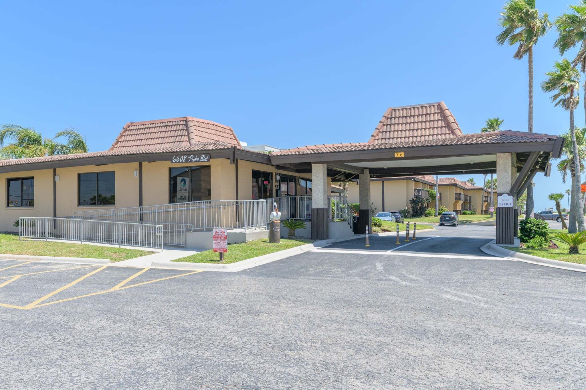 A hotel entrance with a covered walkway, palm trees, and a parking lot.