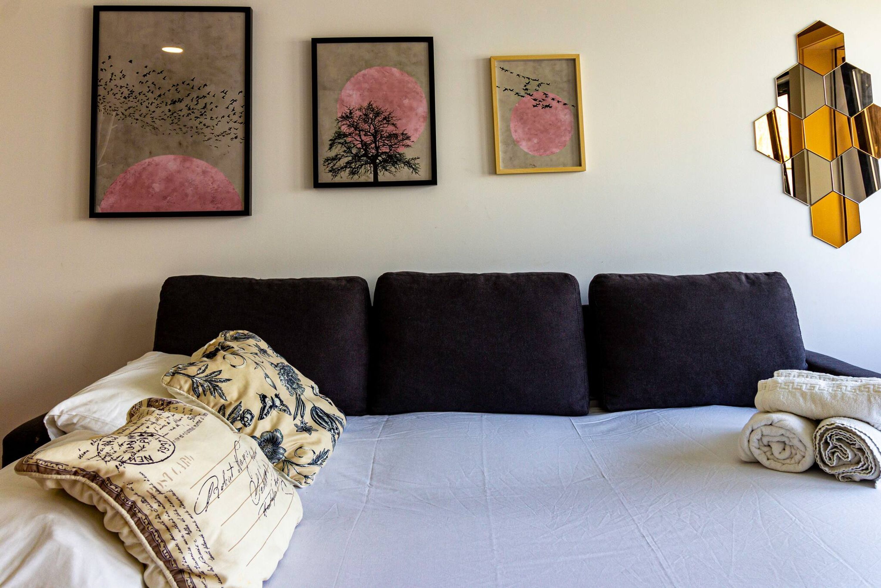 A neatly made bed with dark bedding and decorative pillows, a wall with three framed artworks, and a geometric mirror piece.