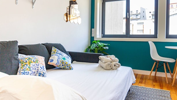 Een moderne slaapkamer met een bed, een klein tafeltje, een stoel, een raam met uitzicht op gebouwen en een wandlamp.