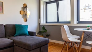 Een moderne woonkamer met een grijze hoekbank, een witte eettafel met stoelen, een groot raam met zonwering, en een decoratief wandobject.