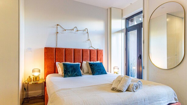 A neatly made bed with a red headboard, two bedside lamps, and a large mirror on the wall. - Guesth4u Heroismo Garden Delight (Porto)