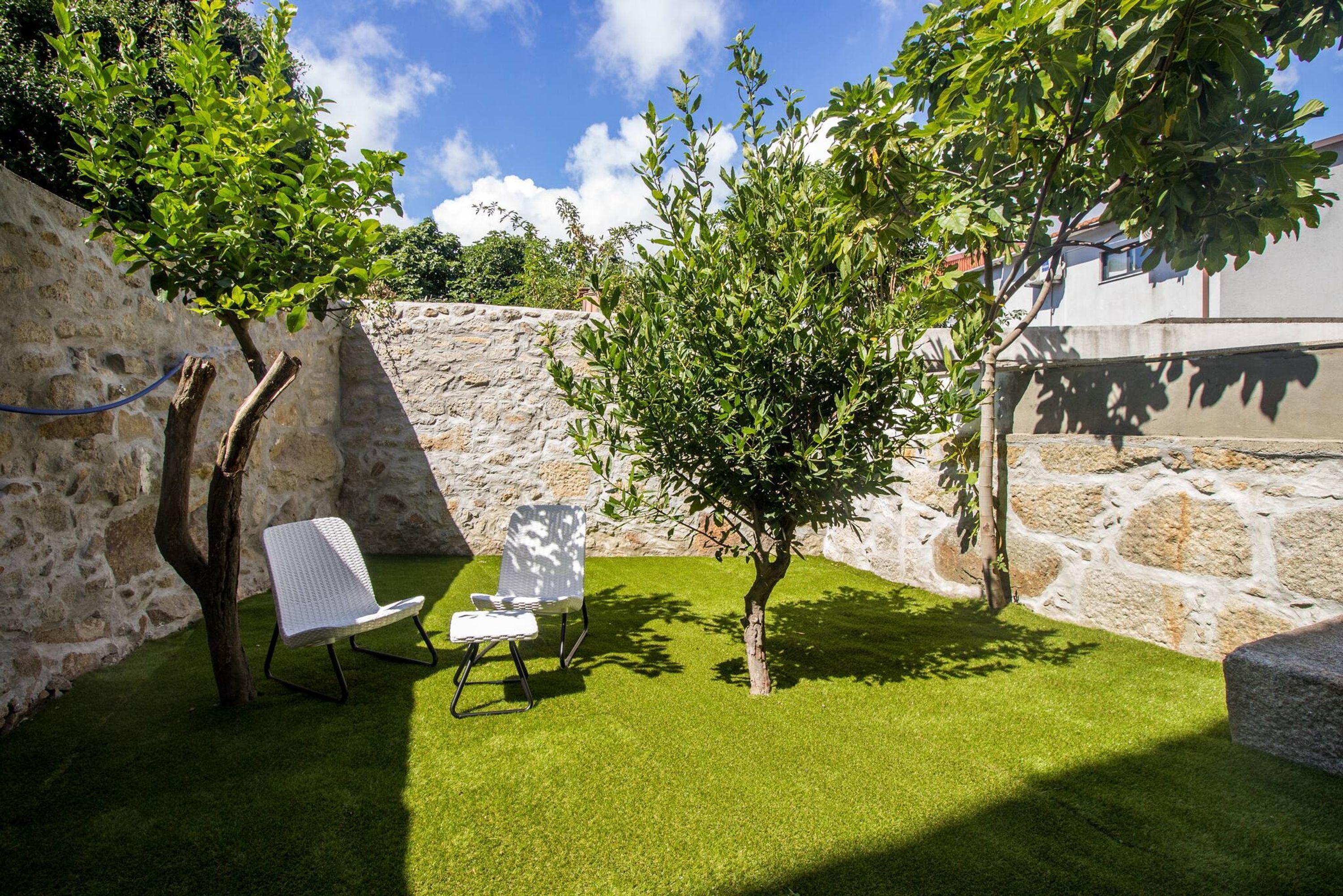 Un mur de pierre, deux arbres et deux chaises blanches dans une cour.
