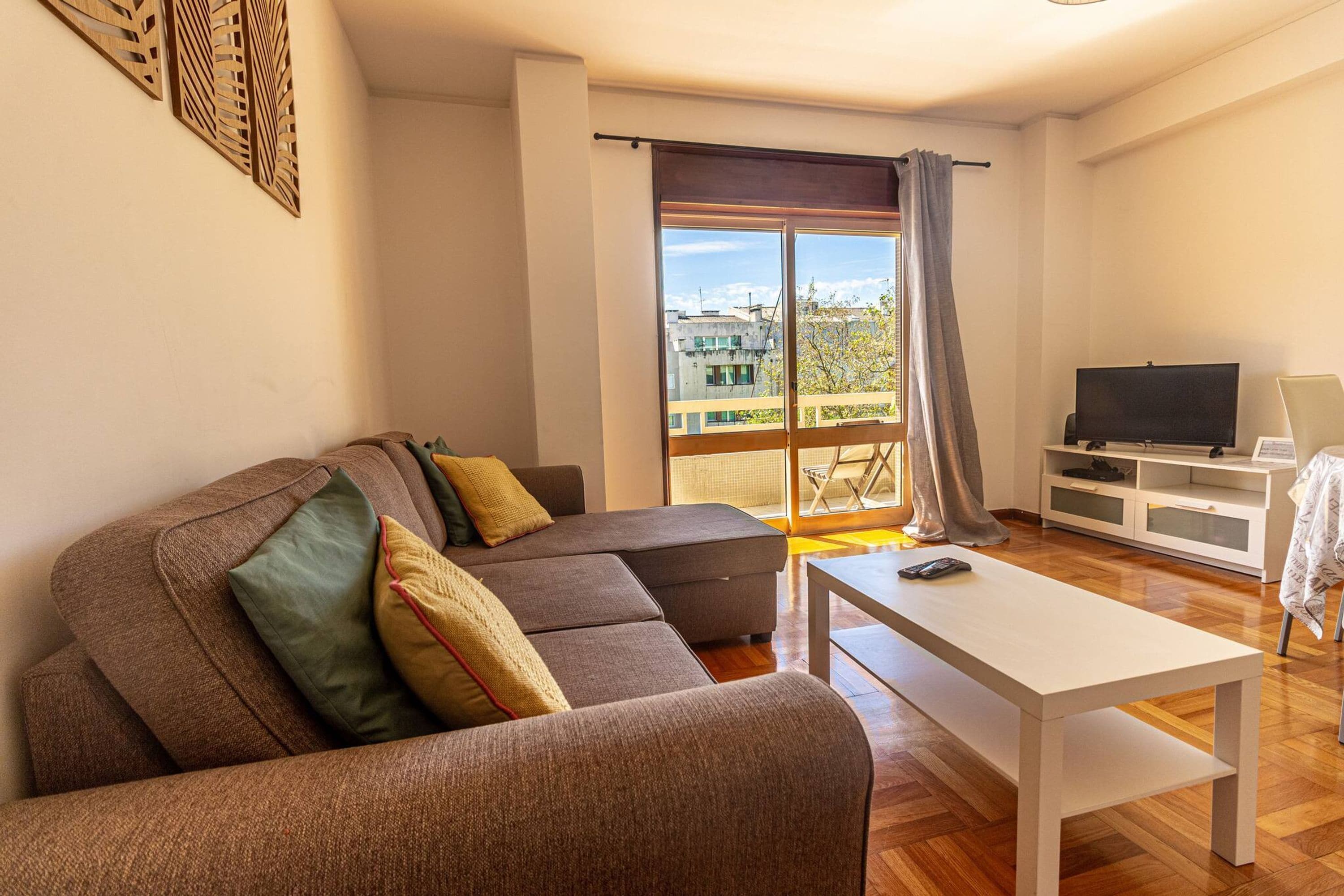 A modern living room with a sectional sofa, a white coffee table, a TV, and a balcony with a view of buildings.