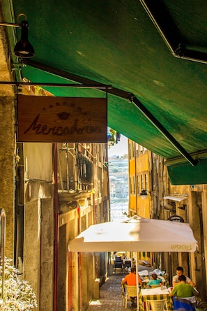 A narrow street with outdoor seating under a green awning, a sign reading "Mercado de", and buildings on either side. - Guest H4U - Porto Ribeira View Mercadores (Porto)