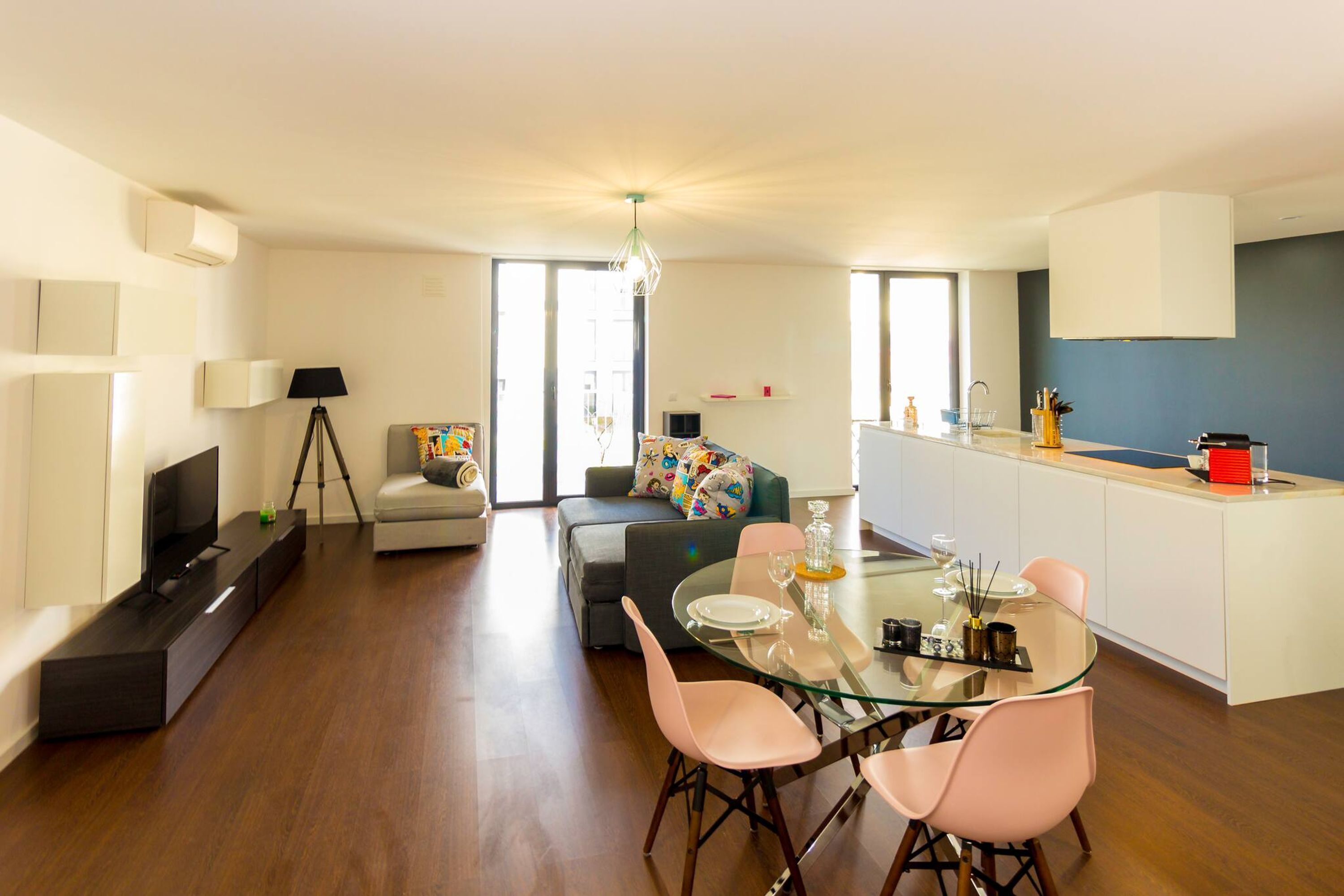 A modern living room with a glass dining table, pink chairs, a sofa, and a TV.