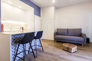 A modern kitchen with a marble countertop and two bar stools. There is a grey sofa and a wooden stool in the living area. - Guest H4U - Hero smo Terrace Ground Floor (Porto)
