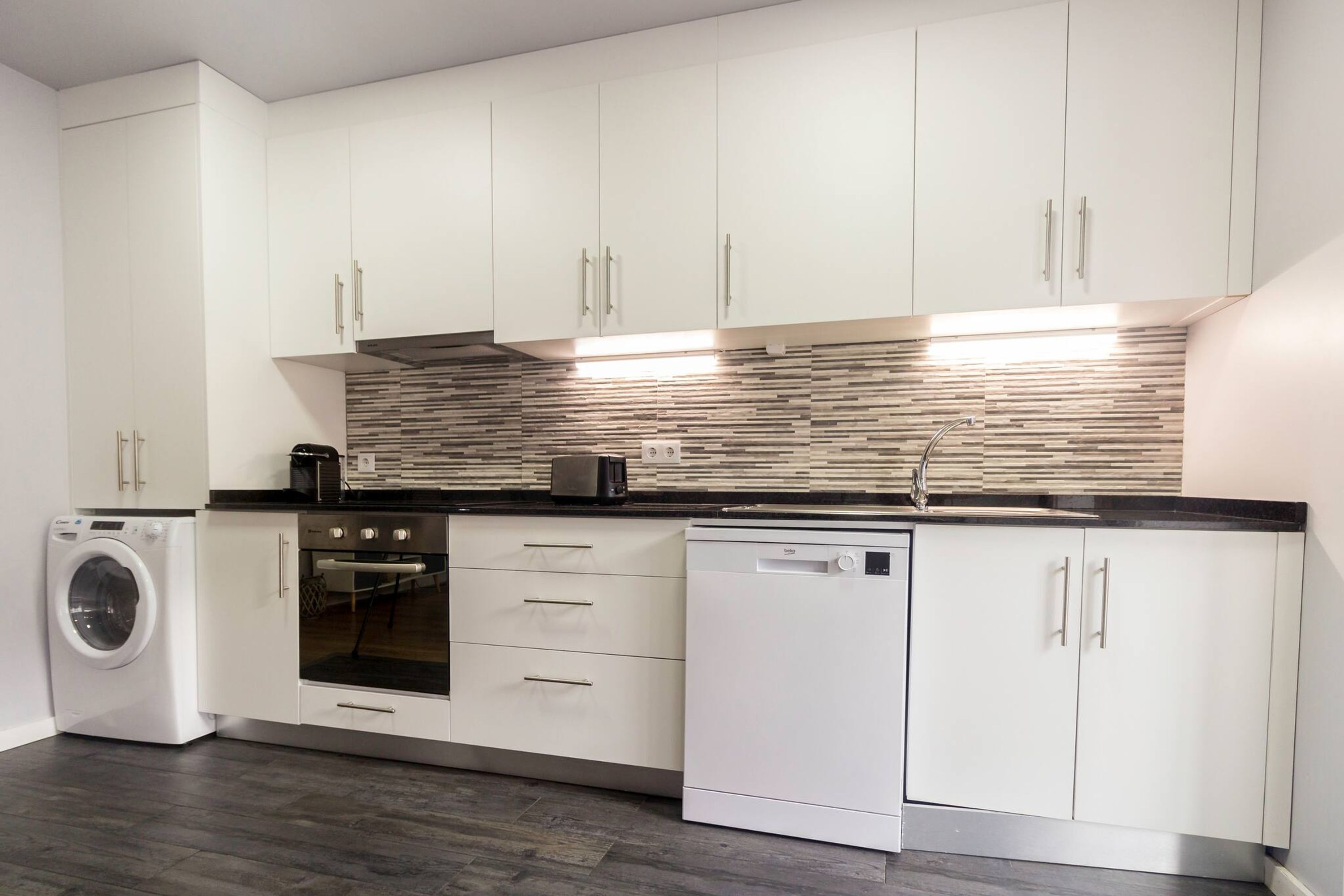A modern kitchen with white cabinets, a built-in oven, a dishwasher, and a washing machine.