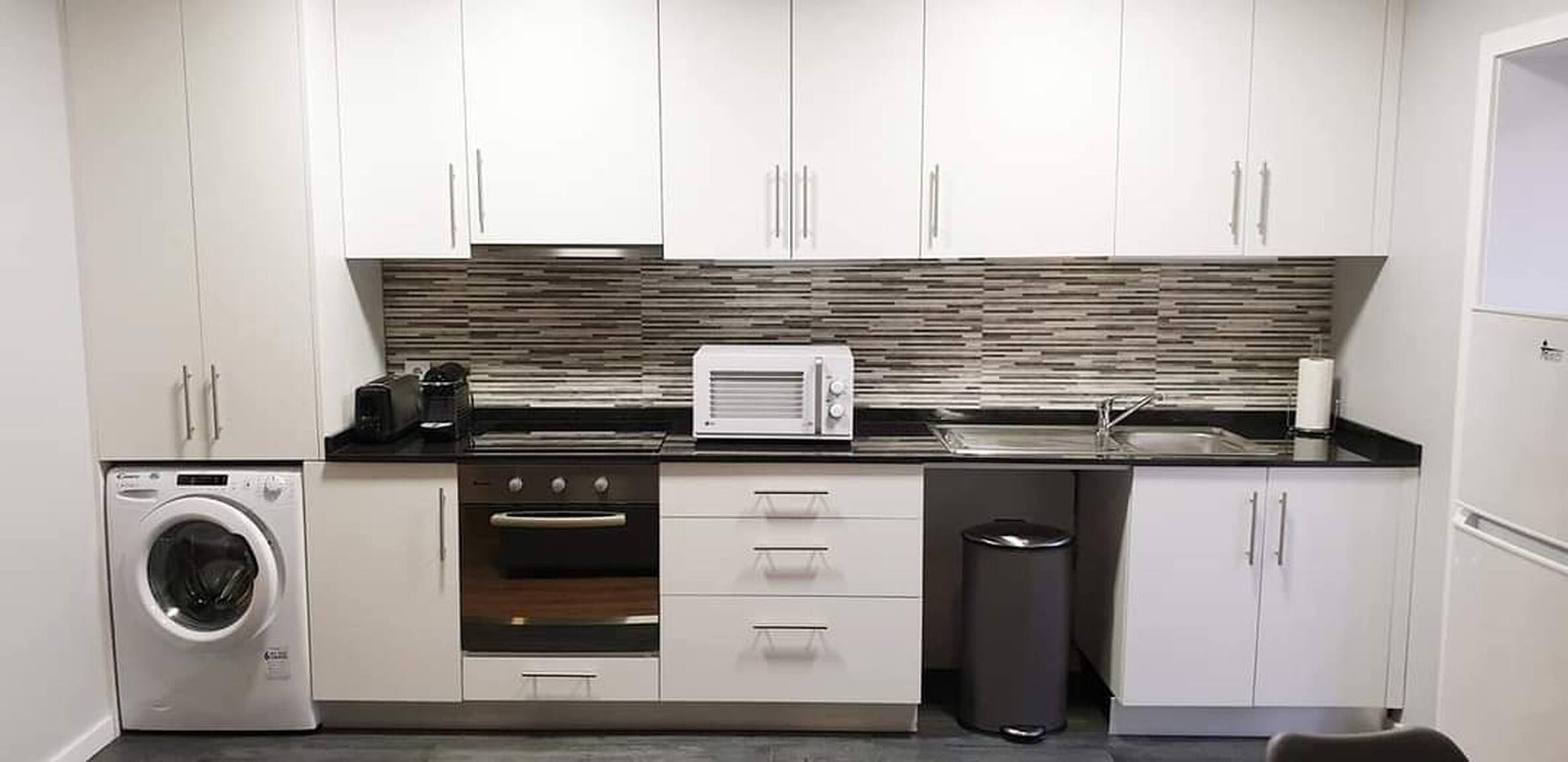 Una cocina compacta con armarios blancos, horno empotrado, microondas y lavadora.