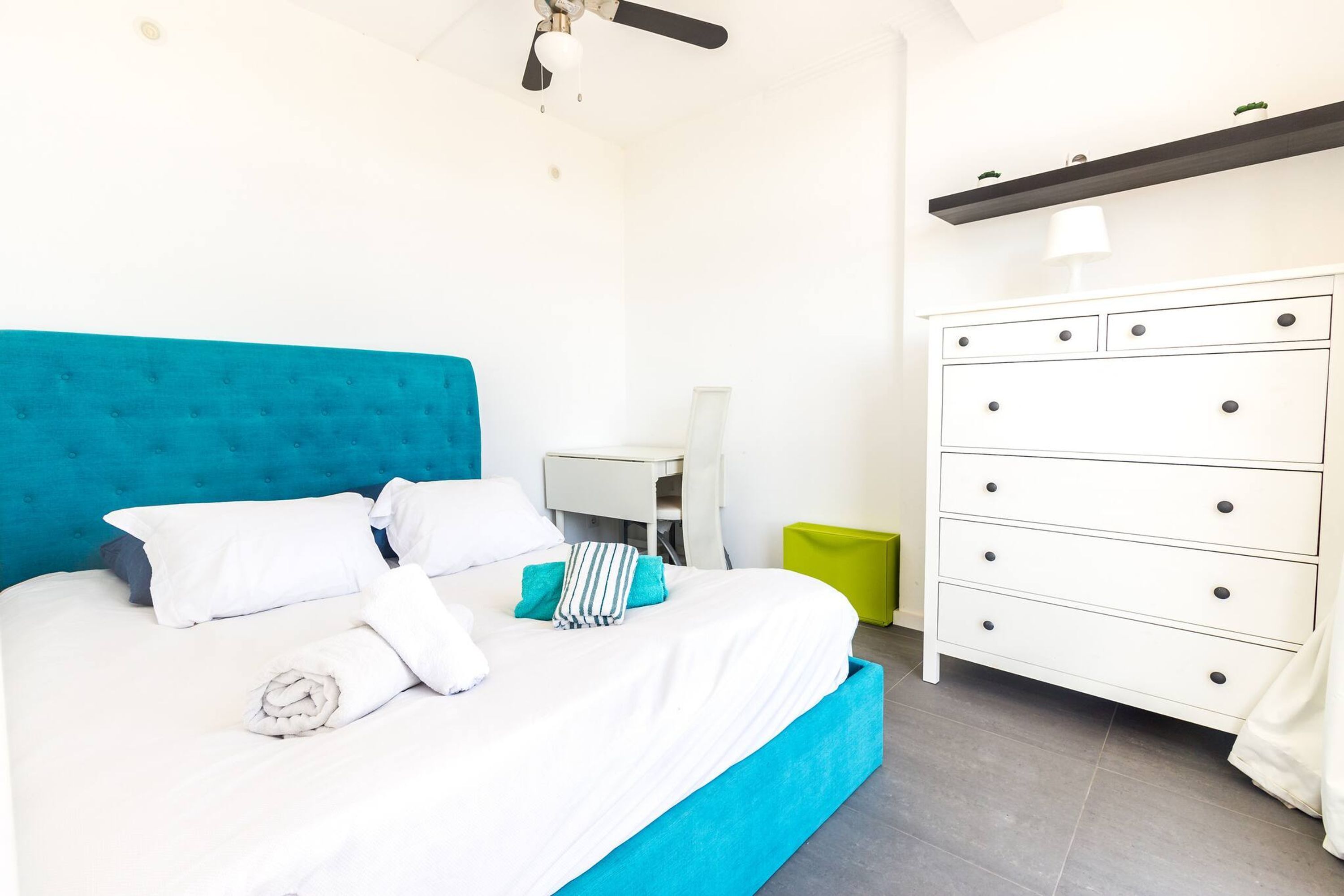 A bed with a blue headboard, white bedding, and a green trash bin.