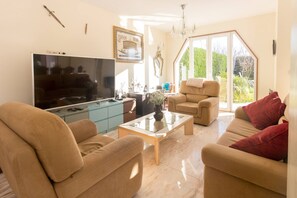 A living room with a large flat-screen TV, beige sofas, a coffee table, and a large window with a view of greenery. - Guest H4U Villa Garden Pool (Povoa De Varzim)