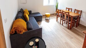 Uma sala de estar moderna com um sofá, uma mesa de jantar com cadeiras, uma mesinha redonda com uma vela e um livro, e uma janela com persianas.