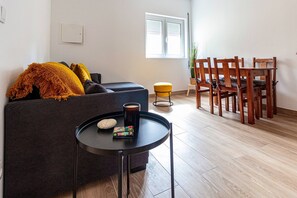 Ein modernes Wohnzimmer mit einer Couch, einem kleinen runden Tisch mit Getränken und Snacks, einem gelben Hocker und einem Essbereich mit Holzsesseln und einem Tisch.
