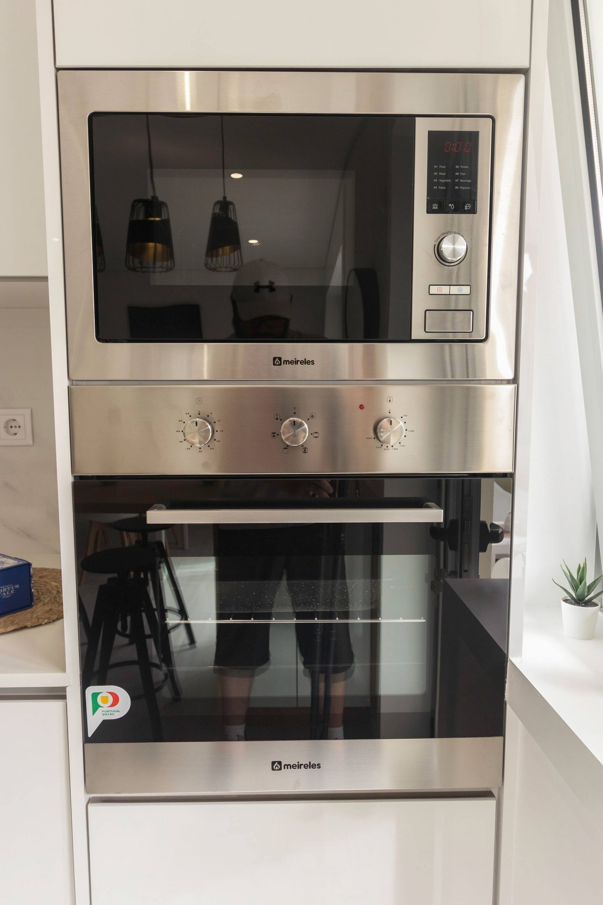 A stainless steel oven with a digital control panel, a lower oven door, and a reflection of a dining area.