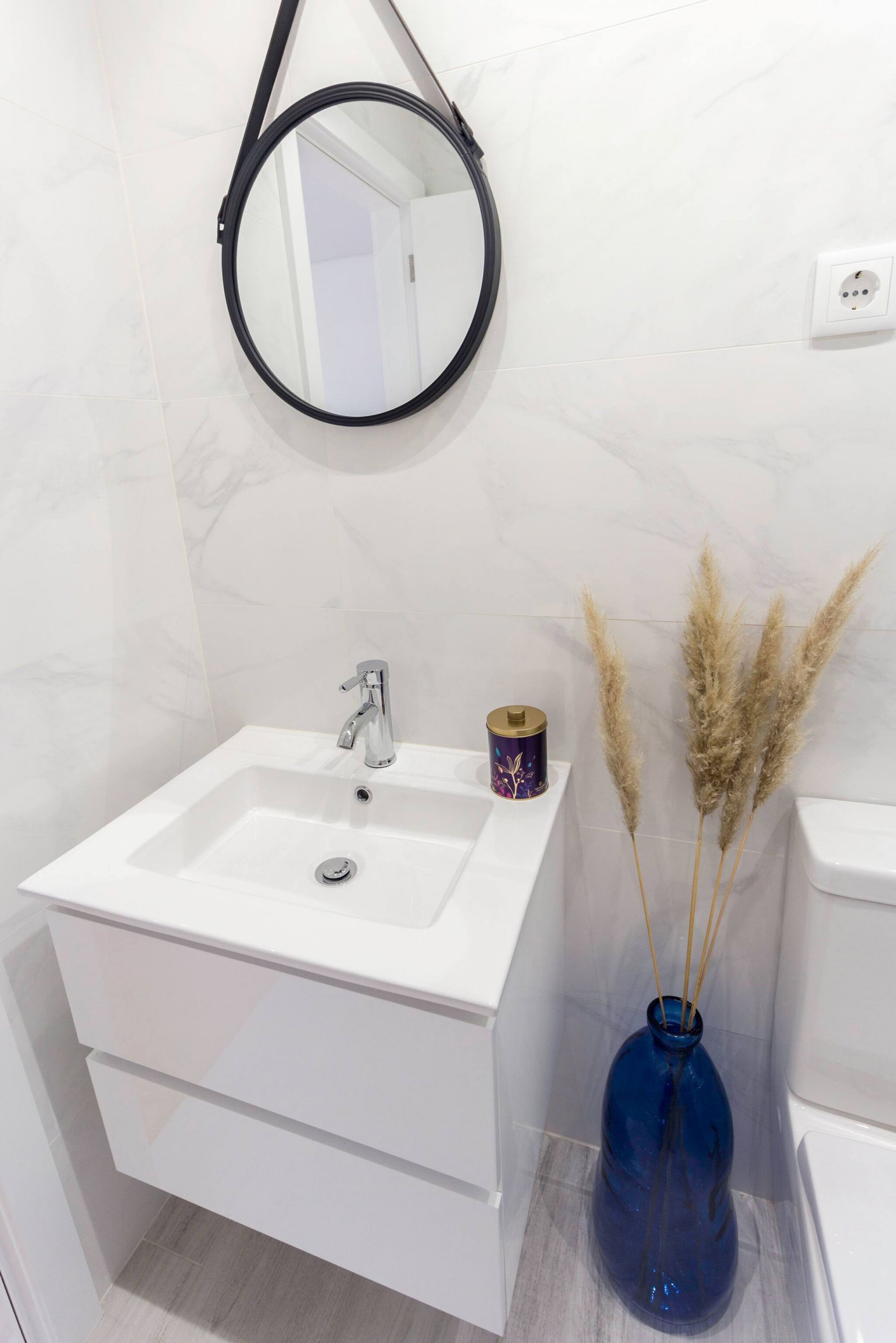 Une salle de bain avec un lavabo blanc, un miroir, un vase bleu contenant des plantes séchées et des toilettes.