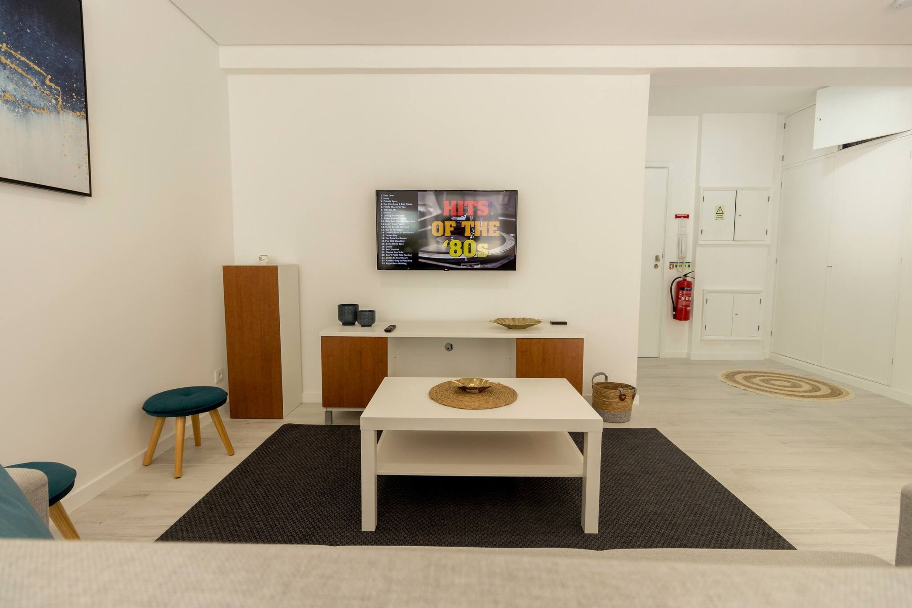 A modern living room with a white sofa, a small round table, and a wall-mounted TV displaying 'HITS OF THE 80s'.