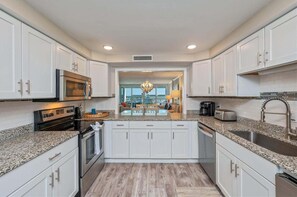 A modern kitchen with white cabinets, granite countertops, stainless steel appliances, and a view to a dining area with a chandelier. - Shoreline Towers 3042 2 Bedroom Condo (Destin)