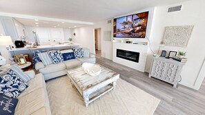 A modern living room with a sofa, coffee table, fireplace, TV, and kitchen in the background. - Shoreline Towers 1044 2 Bedroom Condo (Destin)
