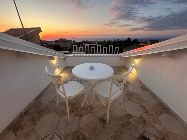 A rooftop terrace with a round table and two chairs, a railing, and a view of the cityscape at sunset. - Godano Rooms (Drapia)