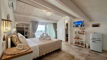 A bedroom with a bed, a chandelier, a television, a refrigerator, and a view of the sea.