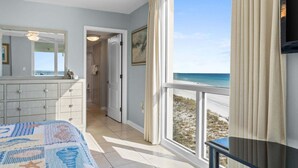 Un dormitorio con un amplio ventanal que da a una playa, una cama con colcha azul y blanca, un aparador y una puerta que conduce a un baño.