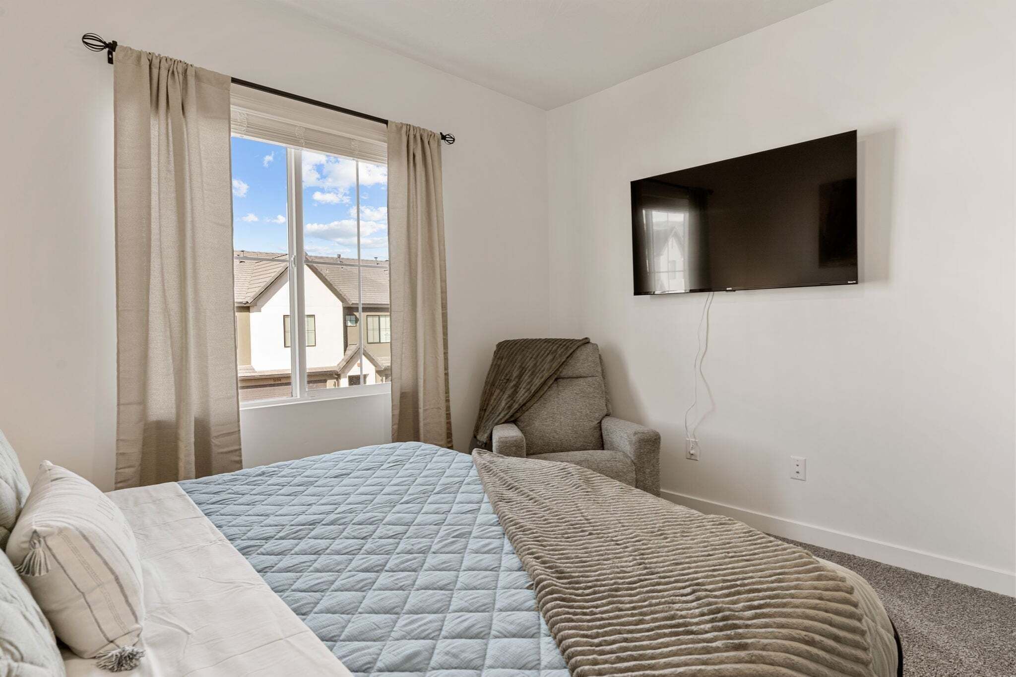 Un dormitorio con cama, una silla, televisión montada en la pared y una ventana con cortinas.