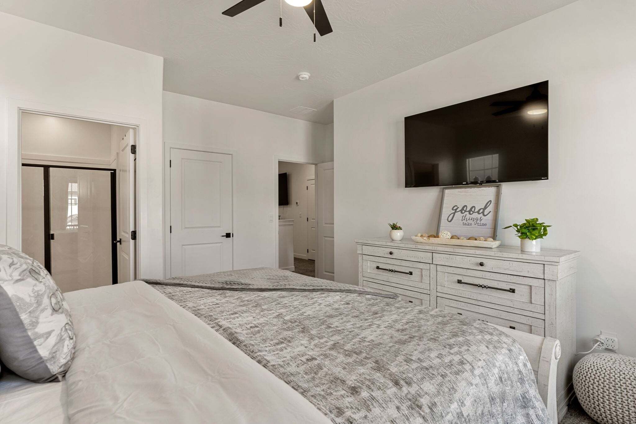 Une chambre à coucher comprenant un lit, une commode, un téléviseur fixé au mur et une porte donnant sur une autre pièce.