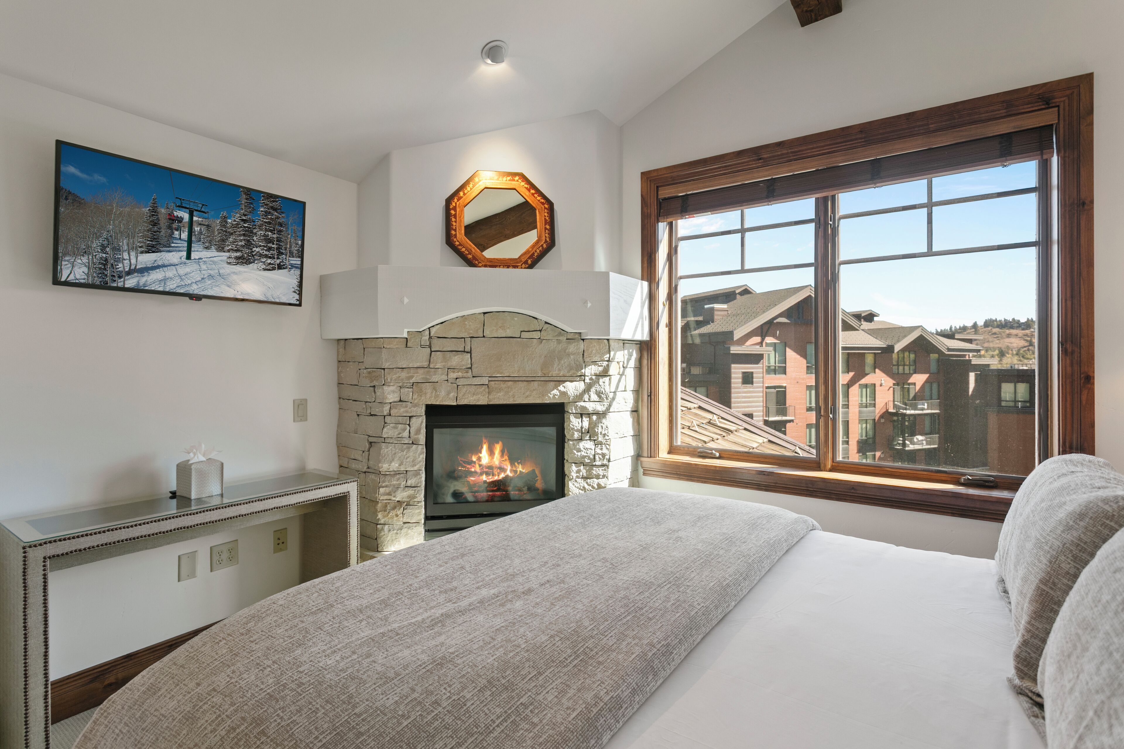Ein Schlafzimmer mit Kamin aus Stein, einem großen Fenster mit Blick auf Gebäude, einem aufgehängten Gemälde und einem Nachttisch mit Taschentuchbox.